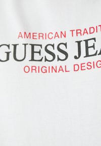 T-shirt blanc avec un texte en gras noir et rouge : "TRADITION AMÉRICAINE DESIGN ORIGINAL GUESS JEANS." Matériau en coton, texture côtelée.