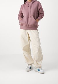 Sudadera con capucha en un tono malva apagado, cuenta con un bolsillo tipo canguro y cordones ajustables. Combinada con pantalones holgados color crema y zapatillas con detalles azules.