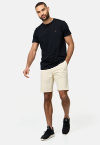 Marineblaues strukturiertes T-Shirt mit kleinem Logo, kombiniert mit cremefarbenen Shorts. Das Modell trägt schwarze Sneakers. Sauberer, lässiger Sommerlook.