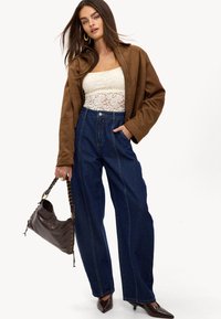 Chaqueta de ante marrón, top de encaje blanco, pantalones vaqueros de denim oscuro de tiro alto y un bolso marrón con cadena. Los tacones de punta completan el look.