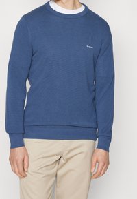 Homme portant un pull bleu texturé à manches longues avec un sous-pull blanc et un pantalon beige, debout devant un fond uni.