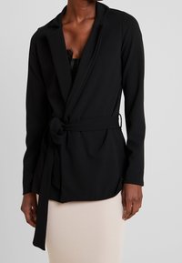 Czarna marynarka o dopasowanym kroju, z kołnierzem w stylu szalowym, korsarzem i tkaninowym paskiem. Gładka tekstura i minimalne zdobienia.