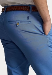 Man som bär ljusblå byxor med en knappad bakficka, brunt flätat bälte och handen i framfickan.