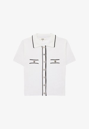 Valkoinen lyhythihainen neulecardigan mustalla reunuksella, edessä napit, terävä kaulus ja kaksi napillista rintataskua.