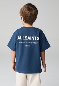 Granatowy bawełniany t-shirt z krótkim rękawem, z dużym białym logo "ALLSAINTS" oraz japońskim napisem na plecach, noszony z jasnymi szortami.