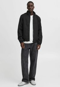 Chaqueta bomber negra con cuello alto, cierre de cremallera y puños acanalados, combinada con pantalones de mezclilla grises sueltos y zapatillas gris claro.