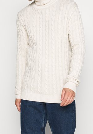 Homme portant un pull à col roulé crème en maille torsadée et un jean bleu foncé, debout devant un fond uni.