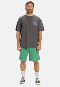Grijze oversized T-shirt met een logo op de borst, gecombineerd met groene corduroy shorts. G gedragen met witte sokken en zwarte sneakers.
