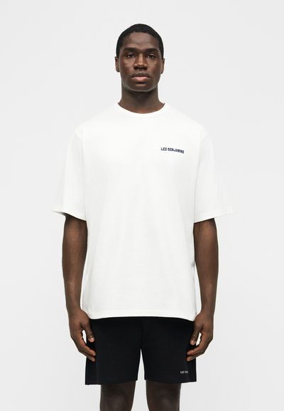 Νέος άνδρας που φοράει λευκό υπερμεγέθη T-shirt και μαύρο σορτς, και τα δύο με μικρά λογότυπα "LES BENJAMINS" στο στήθος και στο μηρό.