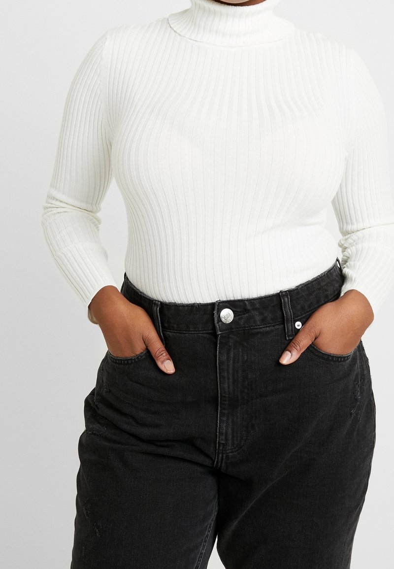 Personne portant un pull à col roulé blanc côtelé rentré dans un jean en denim noir taille haute, les mains dans les poches.
