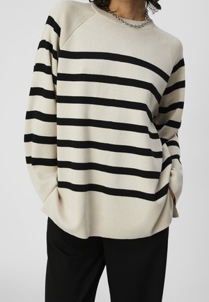 Person trägt einen cremefarbenen Pullover mit schwarzen horizontalen Streifen und eine silberne Halskette, Hände in den Taschen, kombiniert mit schwarzen Hosen.