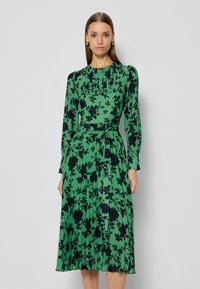 Groene geplooide jurk met een zwart bloemenpatroon, lange mouwen, ronde halslijn en een bijpassende riem in de taille. Glad stofoppervlak.