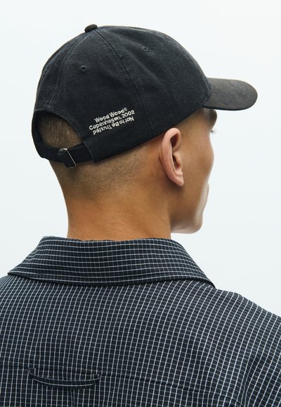 Μαύρο βαμβακερό καπέλο μπέιζμπολ με καμπυλωτό γείσο· διαθέτει κεντημένα γράμματα στο πίσω μέρος. Φοριέται με πουκάμισο σε ασπρόμαυρο καρό.