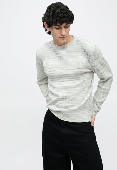 Gaiši pelēks, garām piedurknēm adīts džemperis ar horizontālām svītrainām un teksturētu dizainu, kopā ar melnām biksēm. Brīvs piegriezums un apaļa izgriezuma apkakle.