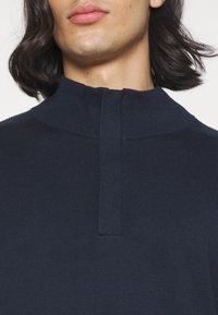 Pull-over en tricot bleu marine avec un col haut et une courte fermeture éclair. Tissu texturé avec des accents côtelés au niveau de l'encolure et des épaules.