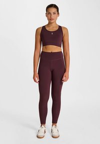 Vinröd sportbehå och högmidjade leggings i stretchigt tyg. Märkbart logodetalj på behån, modellen har vita sneakers.