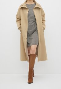 Abrigo exterior beige con un cuello alto y botones dorados, superpuesto a un vestido gris texturizado. La modelo lleva botas marrones hasta la rodilla.