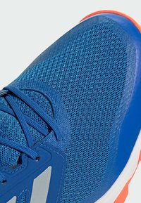 Blå atletisk sko med en andningsbar mesh-överkörning och överliggande detaljer, med en kontrasterande orange sula och grå logotypdetalj på sidan.