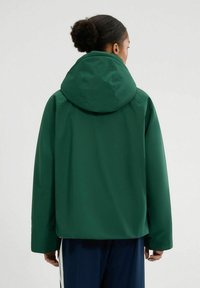 Zielona wodoodporna kurtka z kapturem, gładka faktura, luźny krój i długie rękawy. Widok z tyłu na jasnym tle.