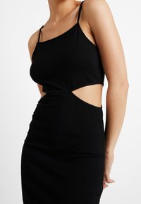 Schwarzes Kleid mit figuroptimierendem Design, ärmellosem Schnitt und seitlicher Ausschnitt-Detail. Hergestellt aus weichem, dehnbarem Material, das eine glatte Textur bietet.