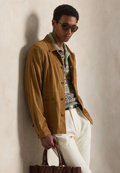 Uomo che indossa occhiali da sole con montatura tartarugata, una giacca marrone, un maglione verde a fantasia, pantaloni bianchi, che tiene una borsa di pelle marrone, appoggiato a una parete beige.