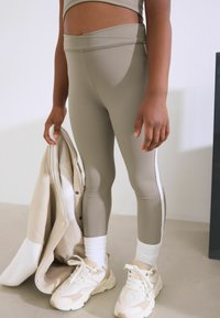 Leggings beiges ajustés avec une taille haute, ornés de rayures blanches sur les côtés. Associés à des chaussettes blanches et des baskets crème, tenant une veste légère.