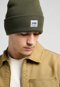 Olijfgroene geribbelde gebreide beanie met een wit rechthoekig logo-patch; gedragen met een tan jas, wat een casual stijl onthult.
