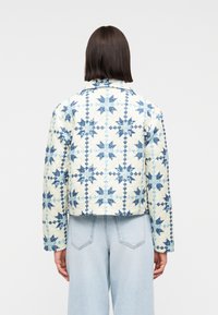 Γυναίκα που φοράει μπουφάν με επένδυση και μοτίβα μπλε και άσπρων αστεριών, συνδυασμένο με ανοιχτόχρωμα τζιν παντελόνια φαρδιάς γραμμής, στραμμένη με την πλάτη στην κάμερα.