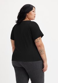 Black short-sleeve t-shirt, relaxed fit, round neckline, smooth texture. Worn with high-waisted dark gray jeans, showing some tattoos on arms.