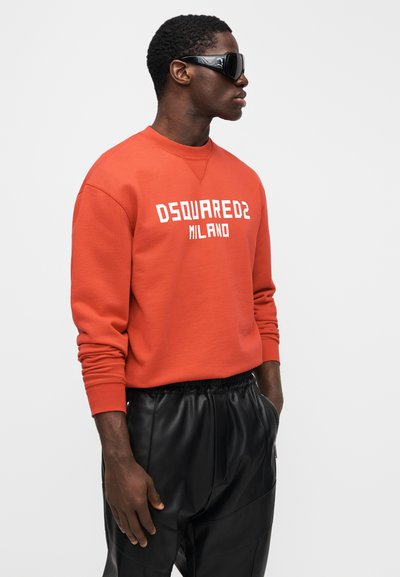 Červená mikina s bílým nápisem „DSQUARED2 MILANO“, kulatý výstřih, žebrované manžety, doplněná černými koženými kalhotami, modelka s brýlemi na slunce.