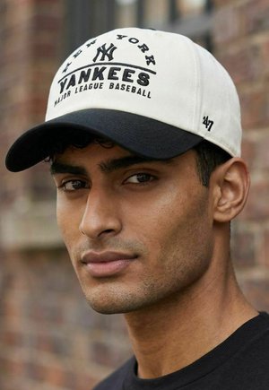 Young man wearing white and black New York Yankees baseball cap, standing in front of a blurred brick wall background.