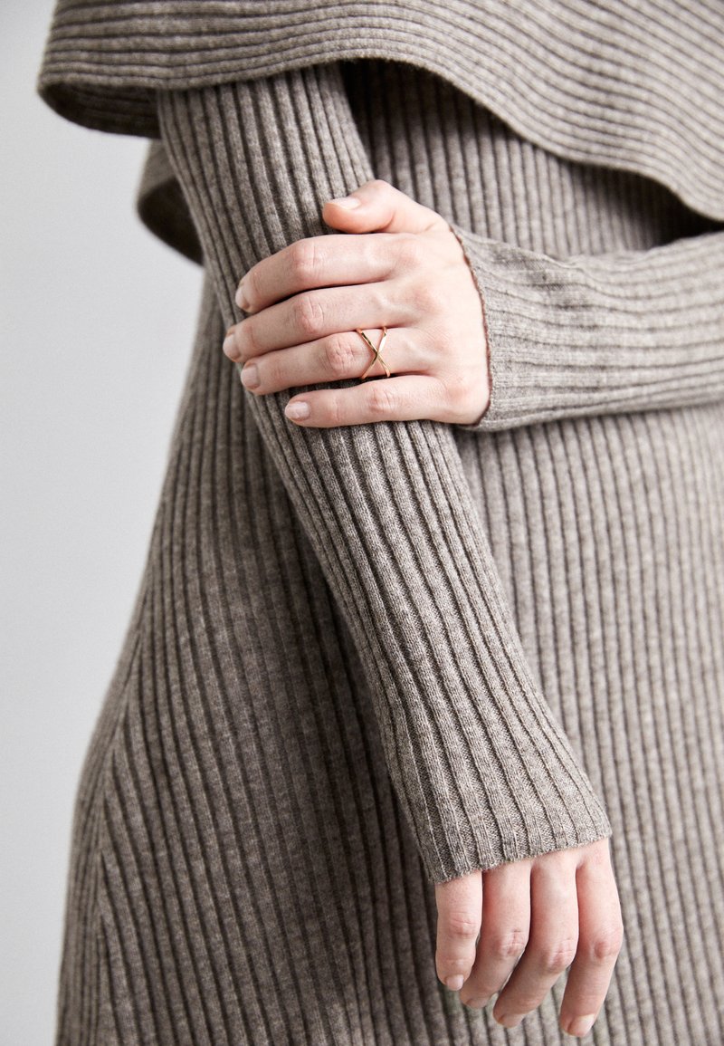 Ribaga, taupe värvi kudum, millel on pikad varrukad. Käsi, millel on kuldne sõrmus, toetub varrukale, näidates selle tekstuuri ja sobivust.