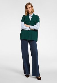 Zielona dzianinowa kamizelka z dekoltem w serek, przednimi kieszeniami i ozdobnym guzikiem, nałożona na jasnoniebieską koszulę. W zestawie z ciemnymi szerokimi spodniami jeansowymi.