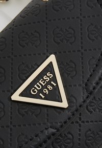 Musta nahkainen käsilaukku, jossa kohokuvioitu kuvio, kultainen kolmionmuotoinen logolaatta, jossa lukee "GUESS 1981", ja ommellut yksityiskohdat reunoilla.