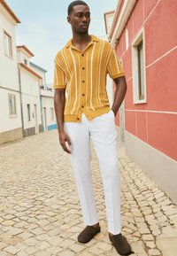 Camisa amarilla de manga corta con textura y rayas blancas, botones negros, combinada con pantalones blancos y zapatos marrones tipo slip-on, de pie sobre adoquines.