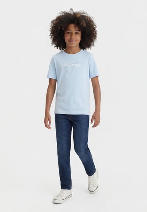 Niño con pelo rizado que lleva una camiseta azul claro de Jack & Jones, vaqueros azul oscuro y zapatillas blancas, de pie sobre un fondo liso.
