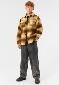 Flanelljacka med gult, brunt och krämfärgat rutmönster, knappstängning, två framfickor, kombinerad med mörka denimbyxor och stövlar.