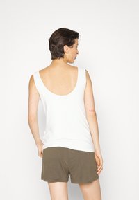 Top branco sem mangas com costas baixas, combinado com calções verdes-oliva. Textura de tecido suave com pele visível nas costas.