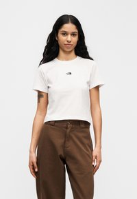 Λευκό βαμβακερό t-shirt με στρογγυλή λαιμόκοψη και κοντά μανίκια, με μικρό μαύρο λογότυπο στο στήθος, σε συνδυασμό με καφέ ψηλόμεσα παντελόνια.