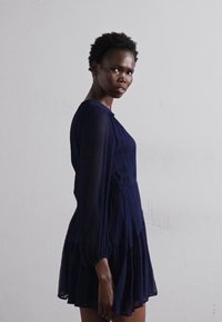 Rochie albastru marin cu mânecă lungă, care prezintă o talie strânsă și o fustă în straturi. Material moale, texturat, cu un luciu subtil.