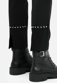 Pantaloni neri con spacchi laterali e dettagli in metallo argentato sull'orlo, abbinati a stivaletti neri in pelle con dettaglio a fibbia.