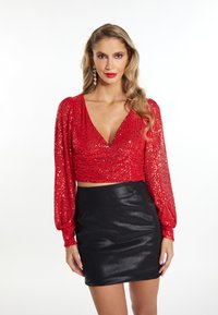 Top corto rosso con paillettes, scollo a V profondo e maniche lunghe a sbuffo, abbinato a una mini gonna nera lucida.