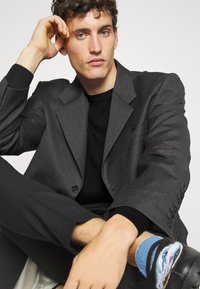 Blazer grigio con revers a punta, chiusura con un bottone e accenti neri. Abbinato a un maglione nero e calzini a fantasia, in posa seduta.