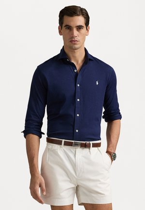 Marineblauwe button-up shirt met lange mouwen, witte knopen en geborduurd logo, gecombineerd met lichtgekleurde shorts en een bruine leren riem.