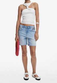 Shorts de mezclilla azul claro con diseños florales bordados en los bolsillos, combinados con un top halter blanco y zapatos de eslora bicolor. Sosteniendo un bolso de cuentas.