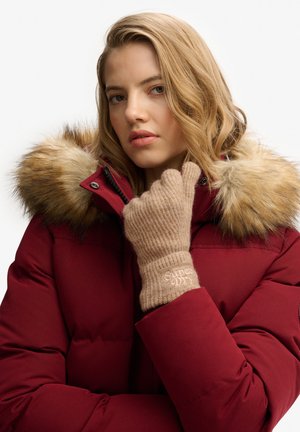 Junge Frau trägt einen roten Wintermantel mit Pelzkapuze, beige Strickhandschuhe und berührt ihr Kinn vor einem einfarbigen weißen Hintergrund.