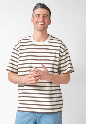 Smaidošs vīrietis ar īsiem tumšiem matiem, valkājot brūnu un baltu svītrainu T-kreklu un gaiši zilas džinsas, stāv pret vienkāršu fonu.