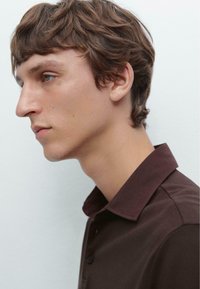  kastanjebruine polo-shirt met een op maat gemaakte kraag, met een gladde textuur en drie knopen. Het zijprofiel van het model is zichtbaar.