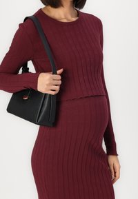 Bordový žebrovaný top s dlouhým rukávem v kombinaci s odpovídající přiléhavou sukní. Modelka drží malou černou kabelku s elegantním designem.