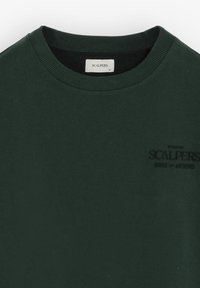 Sudadera de algodón verde oscuro con cuello acanalado. Presenta un logotipo negro bordado en el lado izquierdo. Diseño minimalista, textura suave.